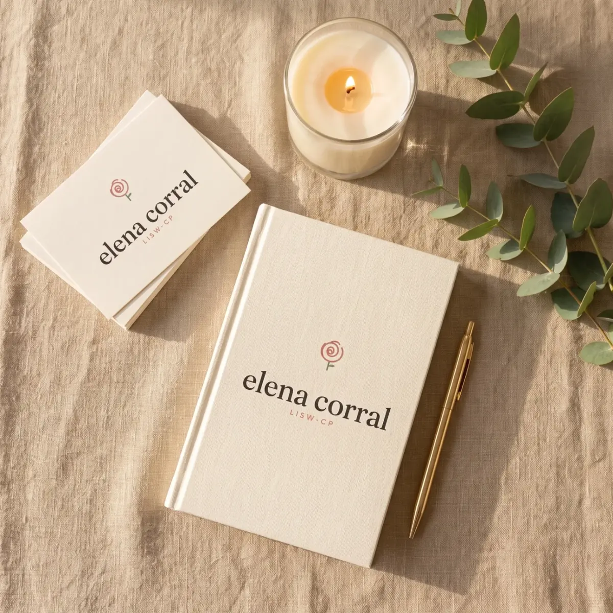 An Elena Corral notebook, business cards, a pen and a lit candle on linen